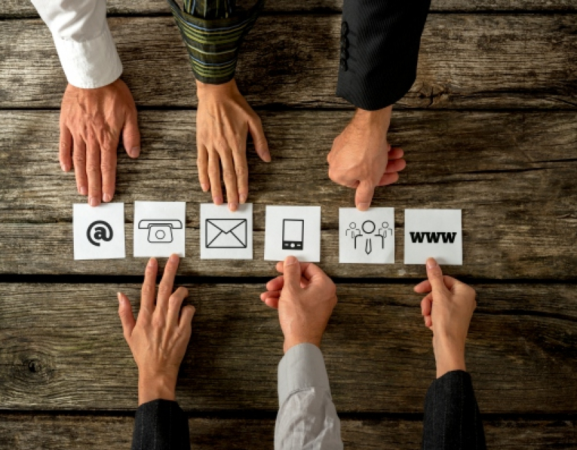 Top view of six people placing white cards with various contact icons in a row on a textured wooden desk.